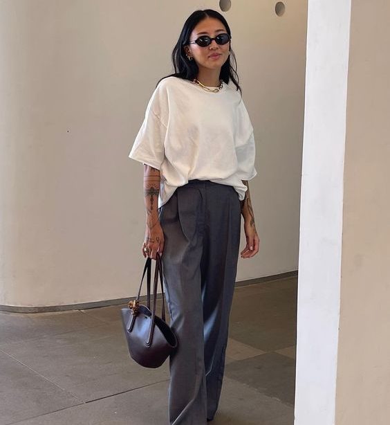 Mulher jovem de pele clara e cabelo escuro. Ela usa óculos de sol e bolsa. Ela veste camiseta branca e calça cinza.