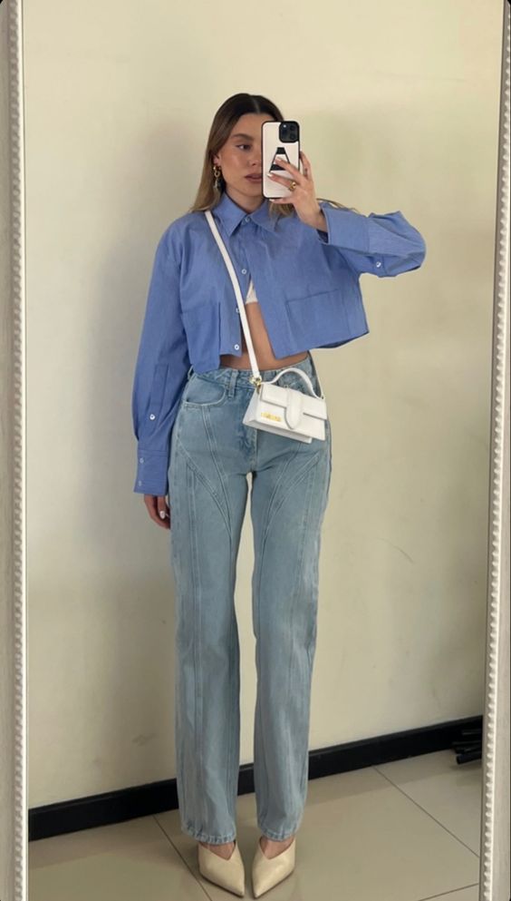Mulher jovem e branca, com cabelo loiro escuro, usando camisa cropped em cor azul, calça jeans, bolsa branca cruzada ao corpo e calçando scarpin.