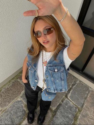 Mulher jovem de pele clara com cabelo loiro. Ela usa camiseta branca e colete jeans.