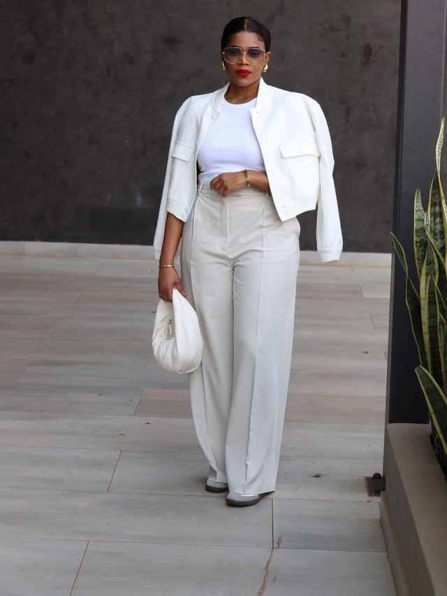 Mulher jovem e negra, com cabelo preso em coque, usando blusa branca, casaco branco, calça de alfaiataria e segurando bolsa meia lua em cor branca.