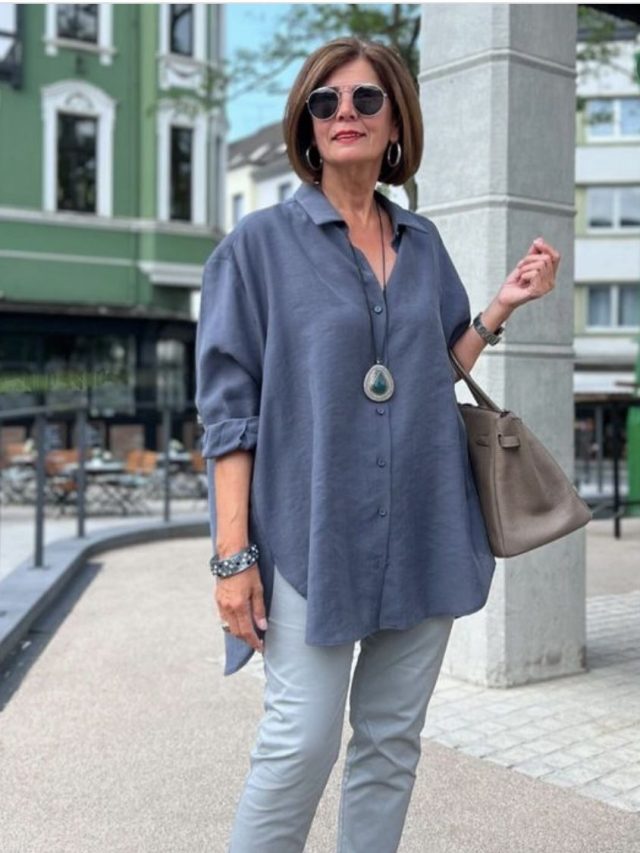 Mulher madura, com cabelo curto e castanho claro, usando óculos de sol, camisa longa em cor azul, calça jeans e sapatilha.