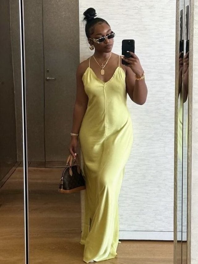 Mulher jovem e negra, com cabelo preso em coque, tirando foto em frente à espelho, usando vestido longo em amarelo-manteiga.