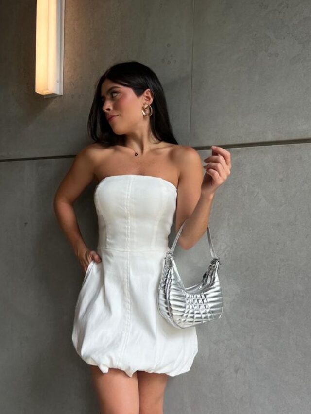 Mulher branca, magra e jovem, com cabelo moreno, usando um vestido balonê sem alça na tonalidade branca e segurando uma bolsa de ombro em cor metalizada.