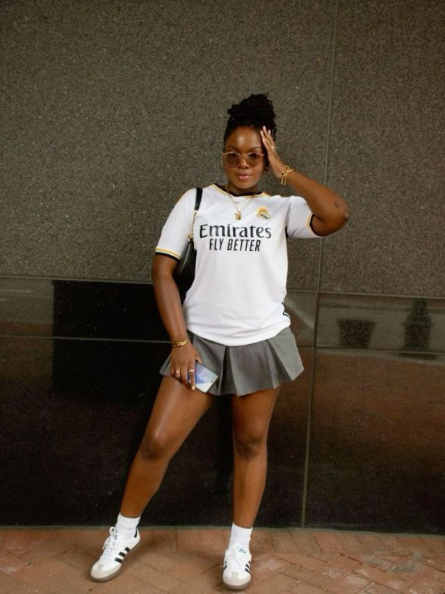 Mulher jovem e negra, com cabelo preso, usando óculos de sol, camisa de futebol, saia de pregas em cor cinza e Adidas Samba.