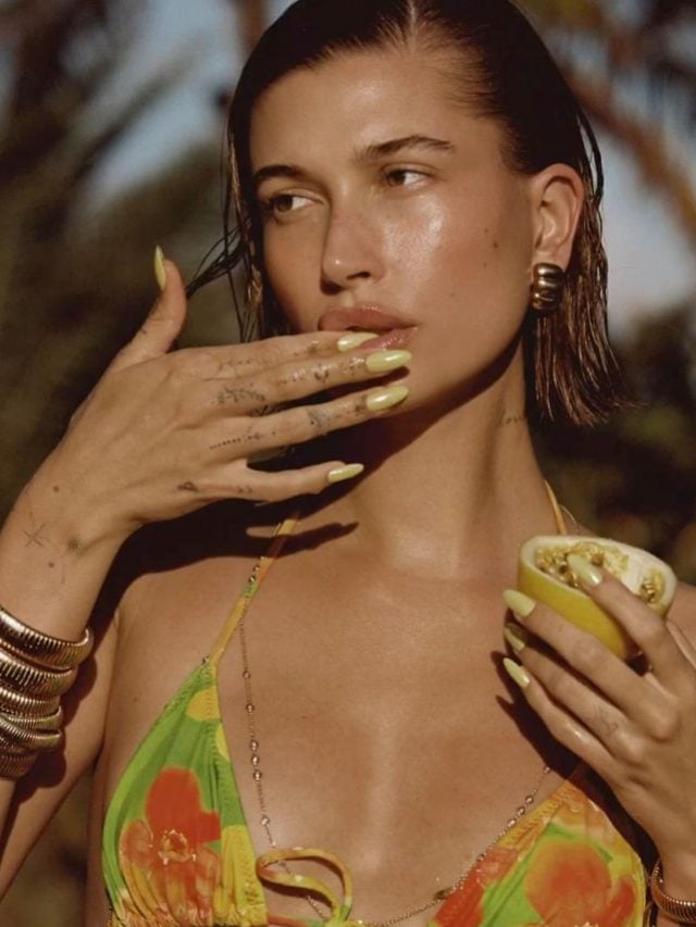 Hailey Bieber com biquini estampado, unha amendoada amarela, segurando um maracujá