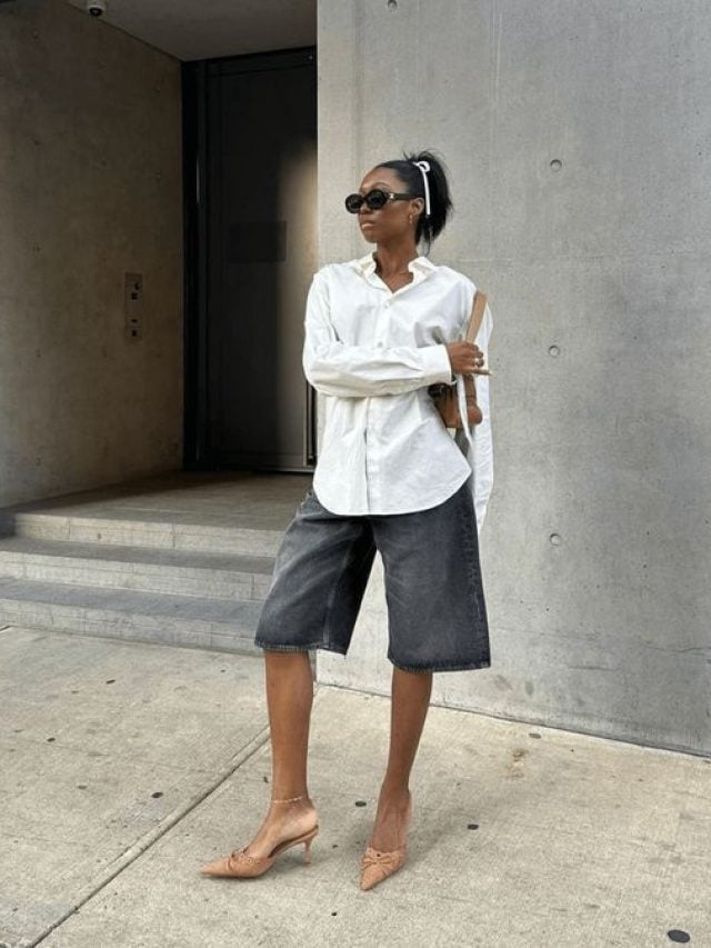 Mulher jovem e negra, com cabelo preso, usando óculos de sol, camisa branca e bermuda jeans.