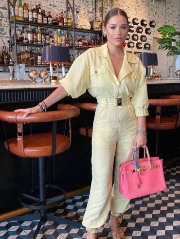 Mulher jovem, branca e com cabelo preso, usando macacão em tonalidade amarelo-manteiga e segurando uma bolsa média em cor rosa.