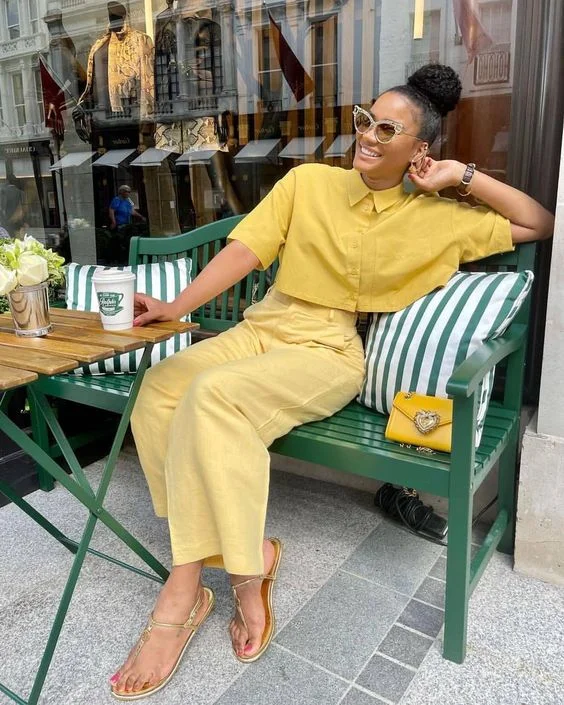 Mulher jovem e negra, com cabelo preso em coque, sentada em banco em frente à mesa, usando camisa cropped de manga curto em amarelo, calça em amarelo-manteiga e sandália rasteirinha.