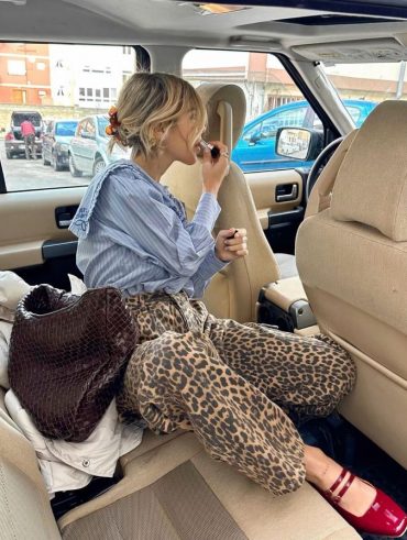 Mulher jovem, loira e branca, com cabelo preso com piranha, sentada dentro de um carro, passando batom, usando camisa azul listrada, calça de oncinha e sapato boneca vermelho.