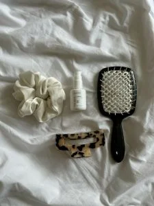 Foto em lençol branco com scrunchie, produto capilar, presilha e escova de cabelo.