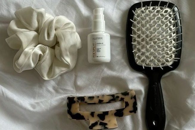 Foto em lençol branco com scrunchie, produto capilar, presilha e escova de cabelo.