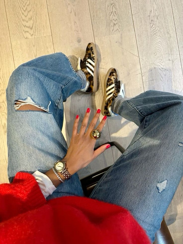 Foto de tronco de uma mulher magra de pele clara. Ela usa Adidas com estampa de oncinha, calça jeans, suéter vermelho, unhas vermelhas e relógio prata. 