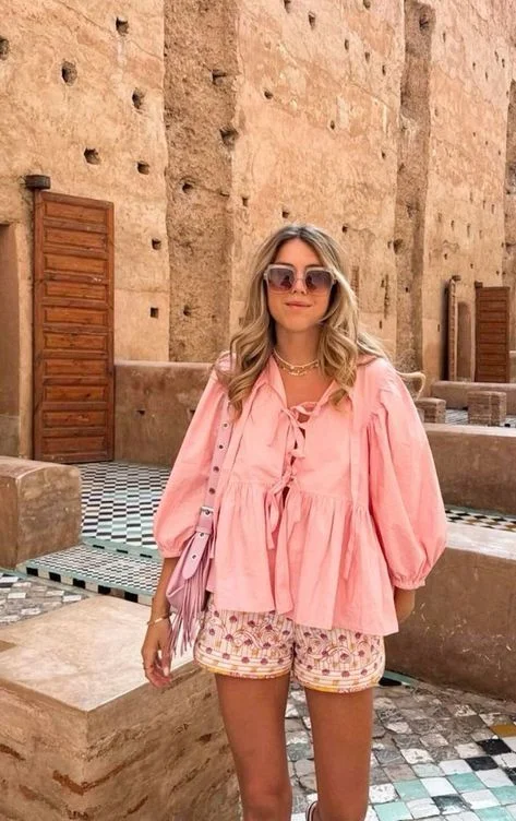 Mulher jovem e loira de cabelo curto, usando óculos de sol, blusa boho com amarações e manga bufante, na tonalidade rosa, com uma bolsa rosa e short rosa.