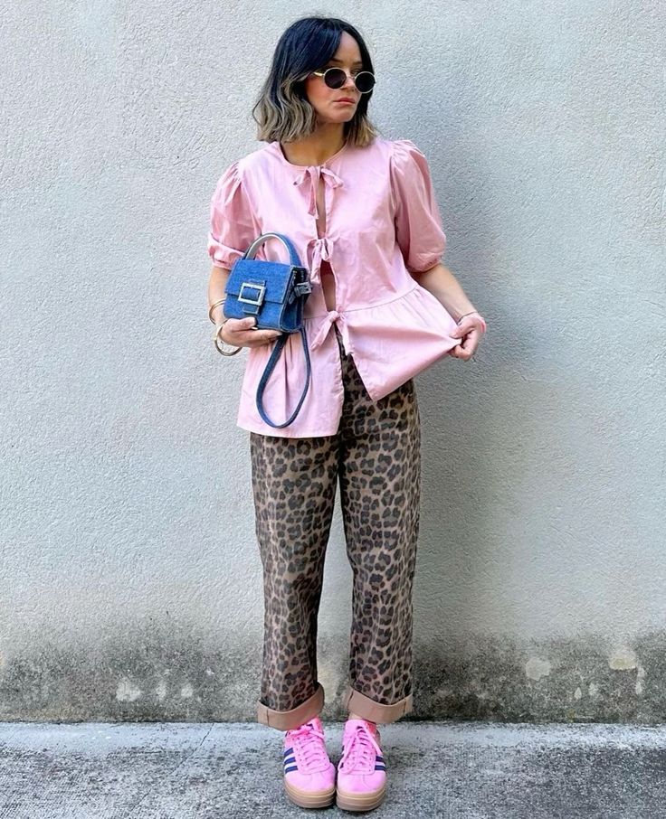 Mulher jovem e branca, com cabelo curto em moreno iluminado, usando blusa boho em cor rosa, segurando uma bolsa com tecido jeans, usando calça de oncinha e tênis Adidas.