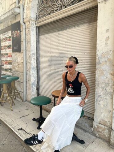Mulher jovem e magra usando saia midi branca, sapatilha preta transparente e colete vermelho