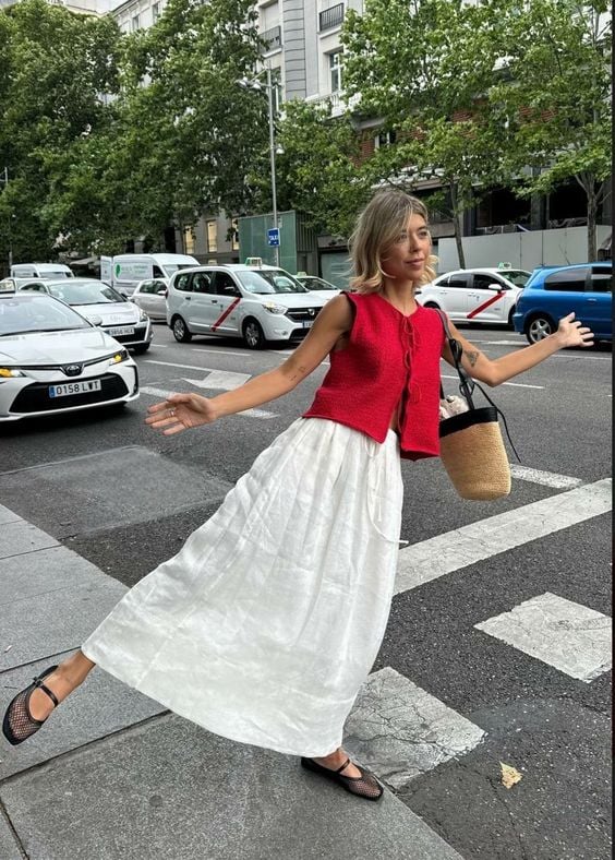 Mulher jovem e magra usando saia midi branca, sapatilha preta transparente e colete vermelho