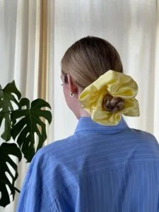 Mulher jovem virada de costas. Ela usa scrunchie amarelo e está de coque. Seu cabelo é loiro claro. Ela veste camisa azul com listras brancas.