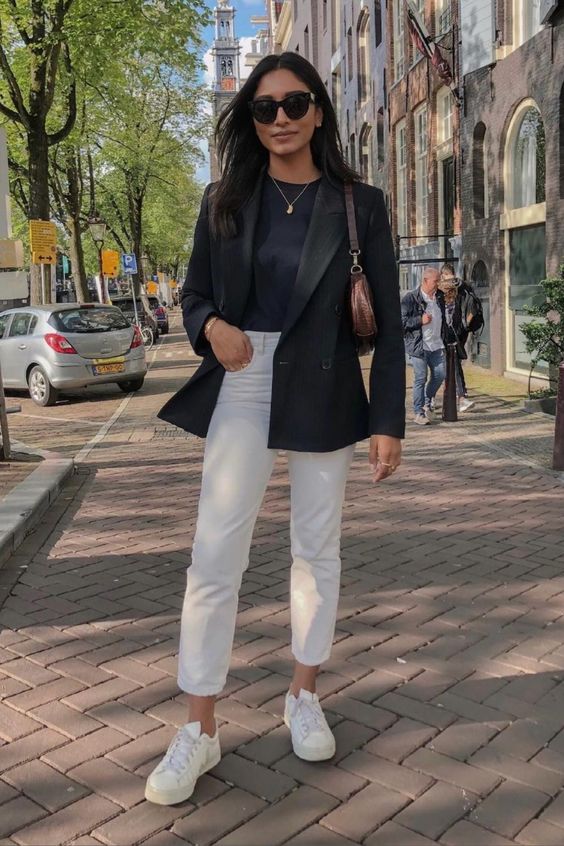 Mulher jovem, magra, de pele clara e cabelo liso escuro. Ela usa blazer preto, calça branca e tênis branco.