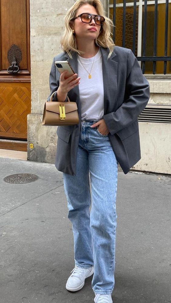 Mulher jovem, magra e de pele clara com cabelos loiros. Ela veste blazer cinza, camiseta branca, calça jeans clara e tênis branco.