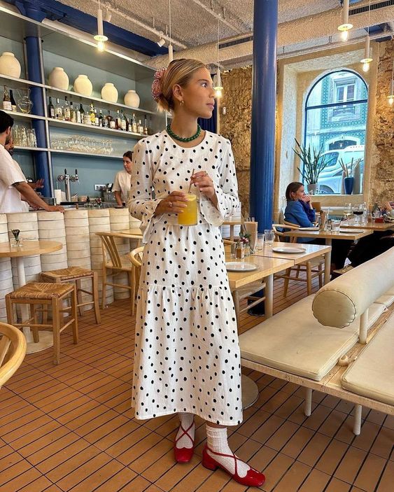 Mulher magra e de pele clara com vestido branco com bolinhas pretas. Ela está usando sapato boneca.