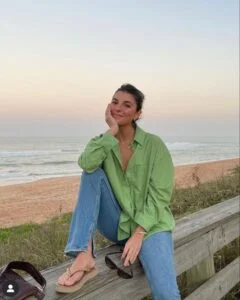 Mulher sentada em estrutura de madeira na beira da praia, usando cabelo preso em coque, camisa verde, calça jeans e chinelo.