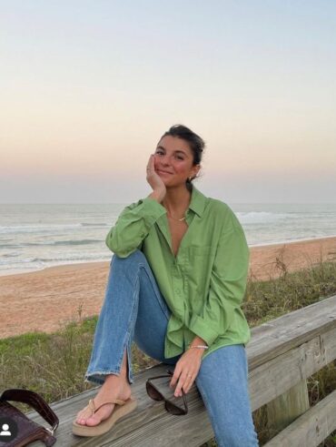 Mulher sentada em estrutura de madeira na beira da praia, usando cabelo preso em coque, camisa verde, calça jeans e chinelo.