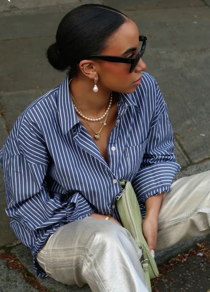 Mulher jovem e negra, com cabelo preso em coque, usando óculos de sol, camisa azul listrada, colar de perolas e segurando uma bolsa pequena em cor verde