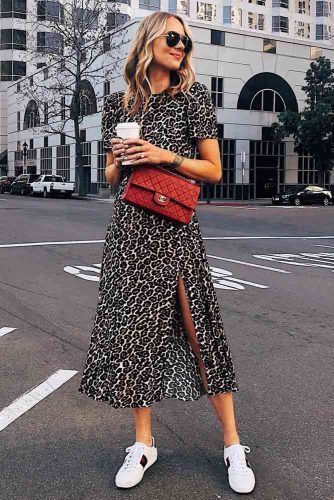 Mulher jovem, branca e loira com cabelo curto, usando óculos de sol, vestido de oncinha, bolsa vermelha, tênis branco e segurando um café