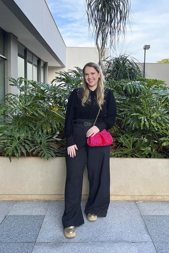 Mulher jovem e loira, usando look preto monocromático, bolsa vermelha e mocassim dourado