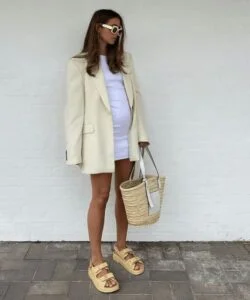 Mulher grávida usando vestido branco com blazer, papete e segurando uma bolsa de palha.