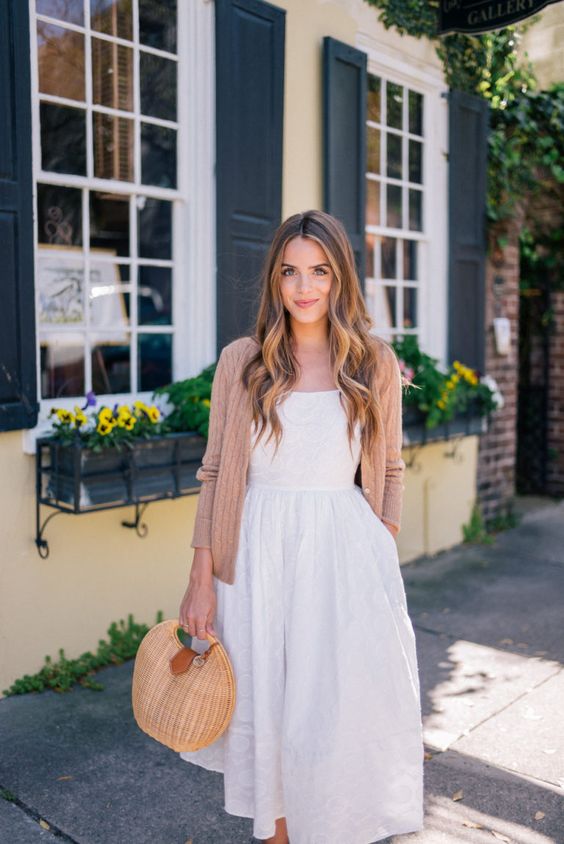Mulher jovem e branca, com cabelo loiro escuro e ondulado, usando cardigan na tonalidade salmão, vestido balonê branco e segurando uma bolsa de palha