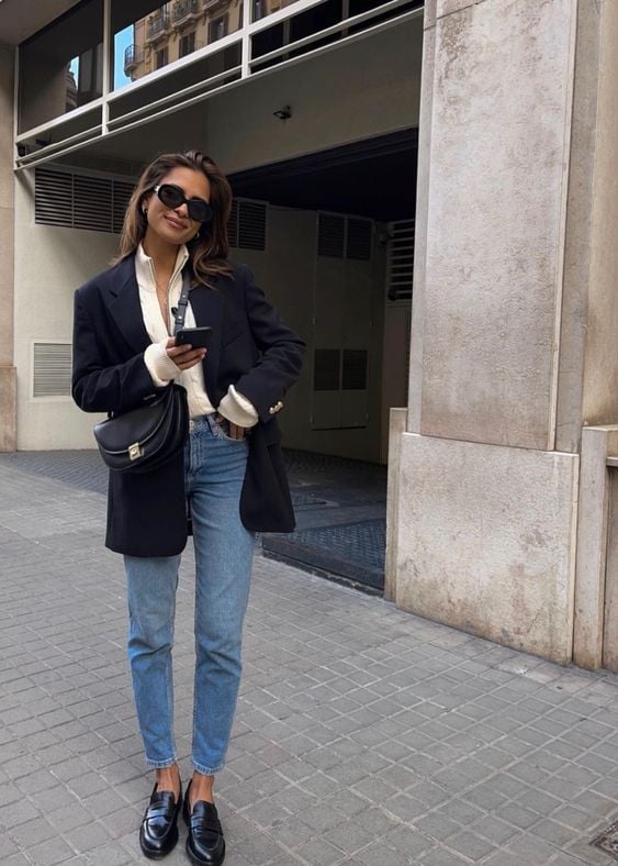 Mulher jovem, magra e branca, usando óculos de sol, camisa branca com blazer preto, calça jeans reta e mocassim
