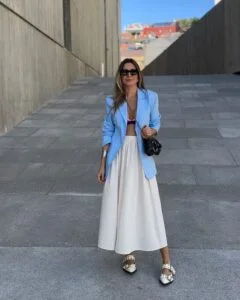 Mulher branca, com cabelo médio em cor castanho claro, usando óculos de sol, blazer azul, saia branca e slingback.