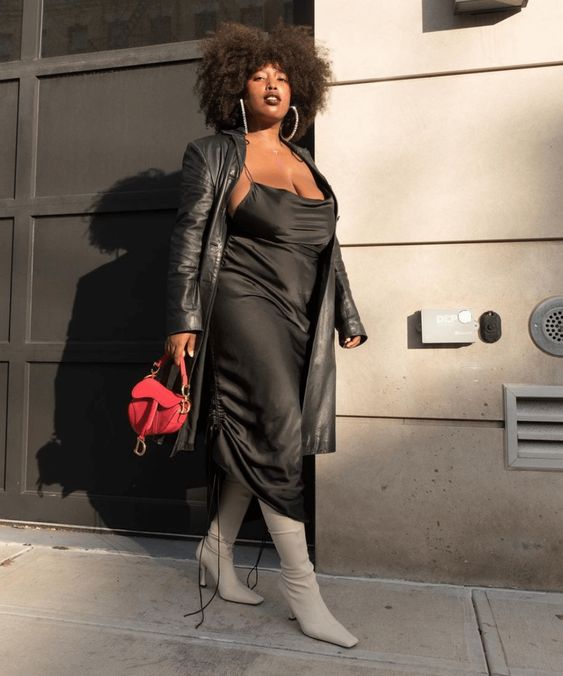 Mulher negra, jovem e plus size, usando sobretudo de couro, vestido de cetim na cor preta, bota de cano alto branca e bolsa vermelha