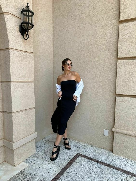 Mulher jovem, branca, com cabelo castanho claro preso em coque, usando óculos de sol, vestido midi em tonalidade preta e sem alça, com camisa branca aparecendo os ombros e uma papete.