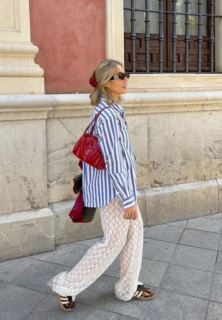 Mulher jovem e loira com cabelo curto, usando camisa listrada, bolsa vermelha de ombro e calça branca de renda