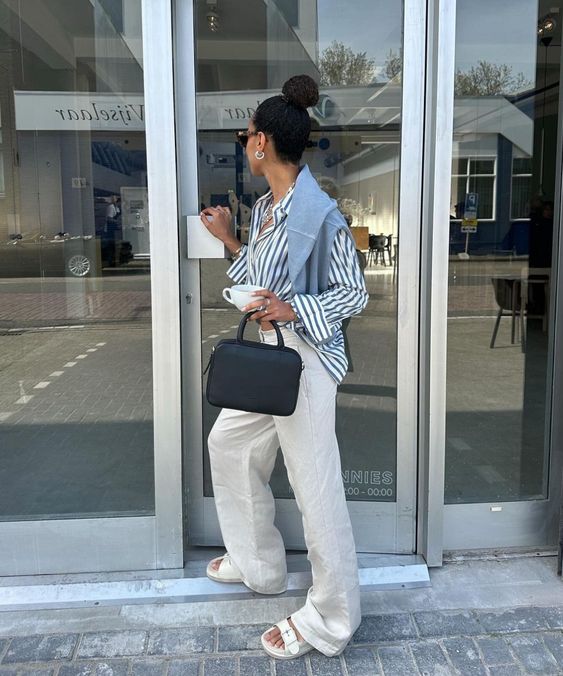 Mulher jovem e com cabelo preso em coque, usando camisa listrada em azul, calça branca, papete e segurando uma bolsa preta