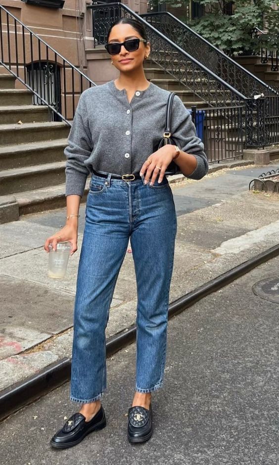 Mulher jovem, magra e branca, usando coque , óculos de sol, cardigan cinza, calça jeans reta com cinto e mocassim