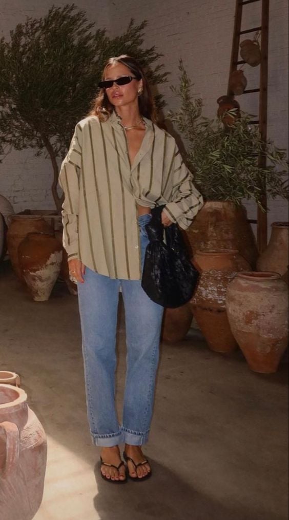 Mulher usando óculos de sol, camisa bege listrada em marrom, calça jeans, chinelo e segurando uma bolsa.