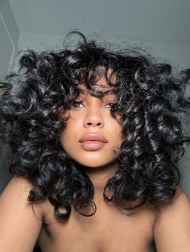 Mulher de pele negra clara com cabelo médio e cacheado, com franja