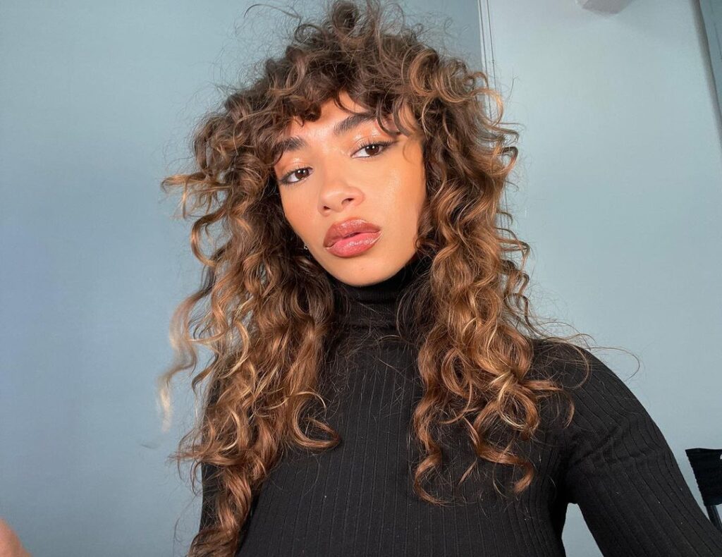 Mulher negra, com cabelo longo, cacheado e castanho iluminado na estética easy chic, com franja e corte shaggy hair