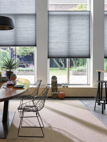 Espaço com mesa oval de madeira, cadeiras de metal e cortinas