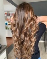 Mulher com cabelo longo e ondulado com babyliss, na cor castanho-escuro iluminado com tom caramelo