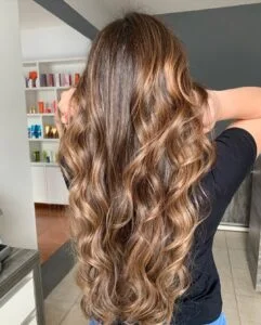 Mulher com cabelo longo e ondulado com babyliss, na cor castanho-escuro iluminado com tom caramelo