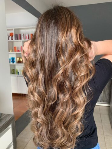 Mulher com cabelo longo e ondulado com babyliss, na cor castanho-escuro iluminado com tom caramelo