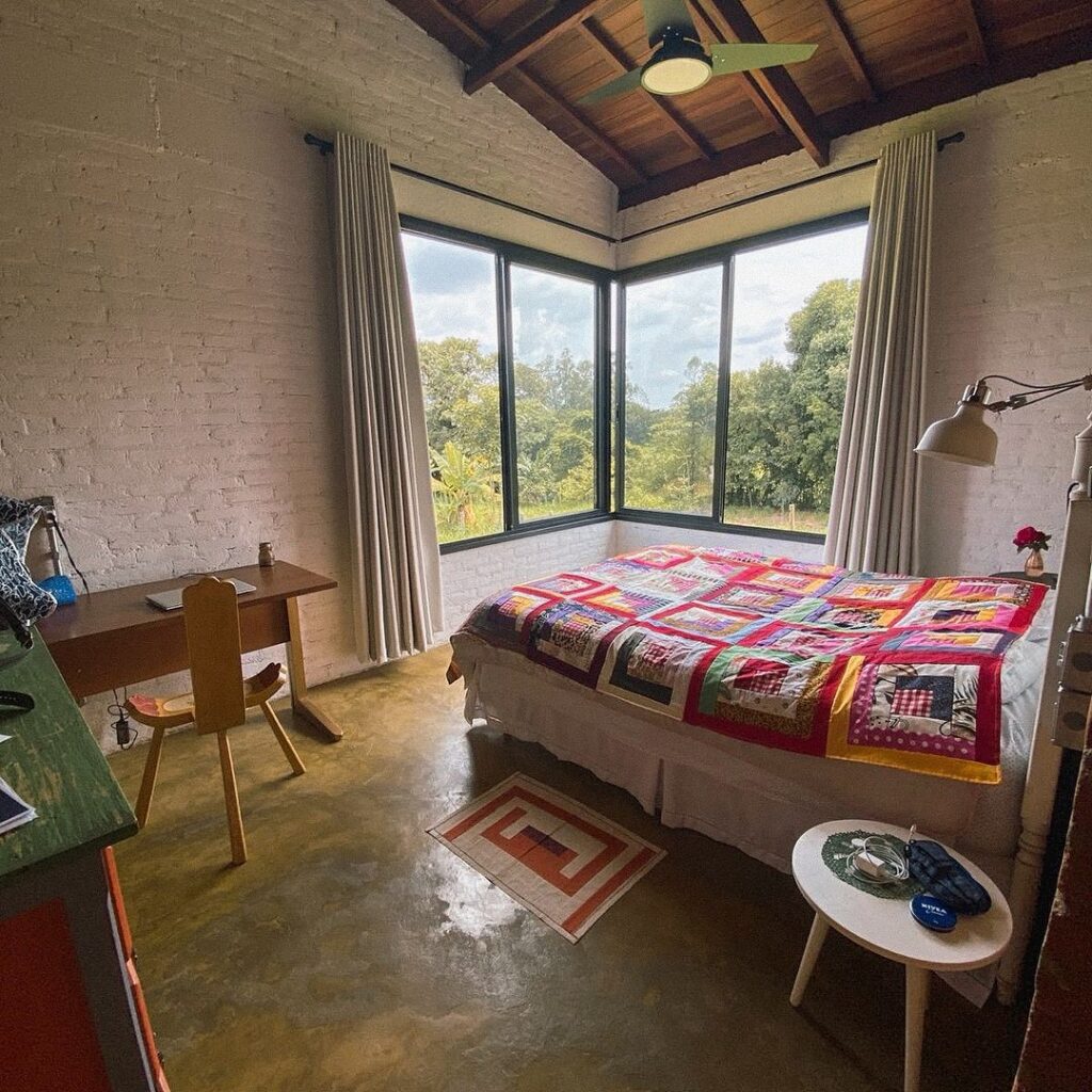 Quarto de casa de campo, com manta colorida, janelas amplas, piso de cimento queimado e parede de tijolos