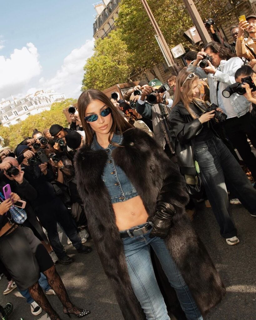 Bianca Andrade na Semana de Moda de Paris, usando sobretudo preto de pelo, luvas pretas de couro, jaqueta jeans e calça jeans de cintura baixa com cinto.