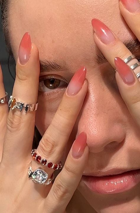 Mulher de pele branca, com as mãos no rosto, com anéis de brilhante e unha amendoada coral ombré 