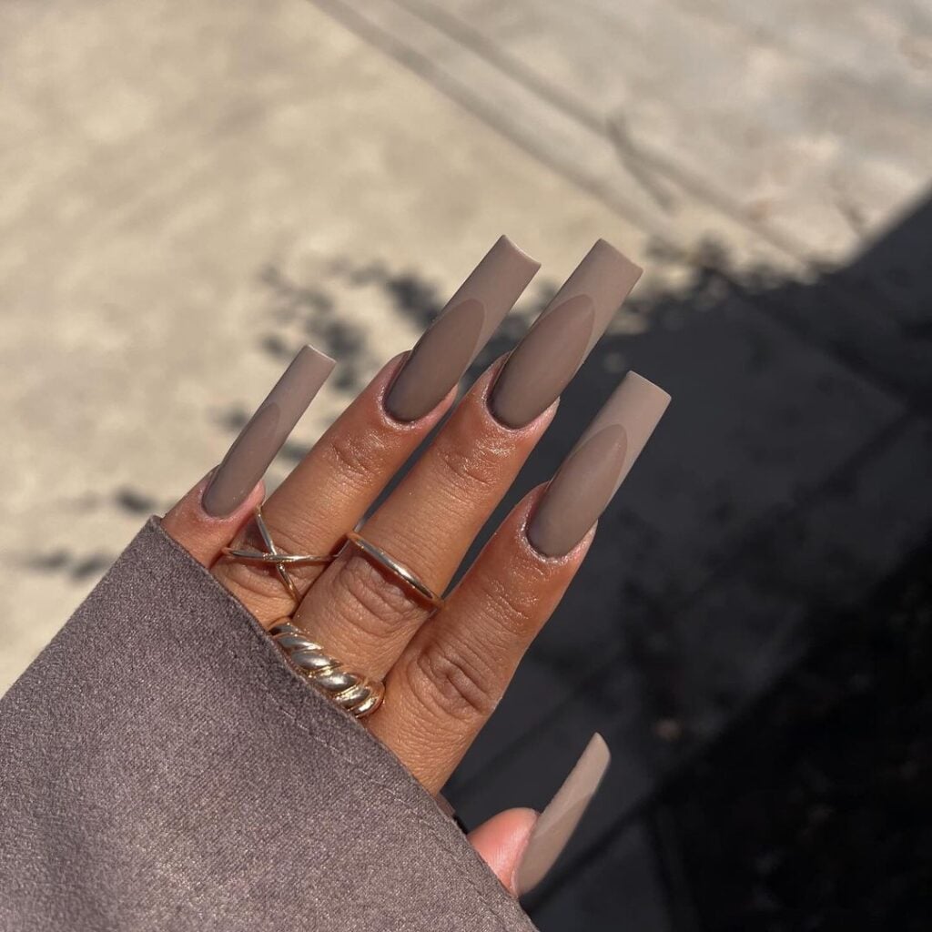 Mão de pele preta, com anéis em prata e dourado e unha comprida e quadrada com esmalte marrom acinzentado
