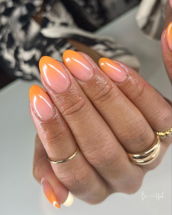 Mão de pele preta, com anéis dourados e unha amendoada ombré laranja 